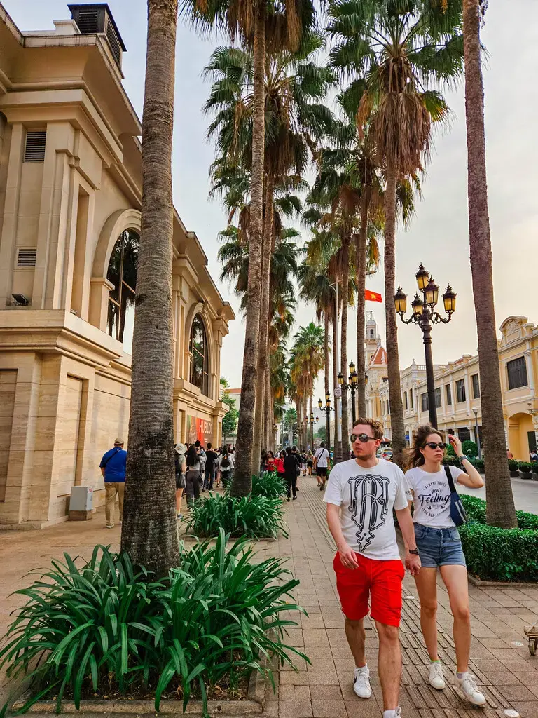 2025胡志明市旅遊指南|從行前準備到在地美食景點,一篇搞懂自由行大小事 胡志明市旅遊步行在街道上,兩旁種植著棕櫚樹,背景有法式殖民風格的建築,行人走在人行道上