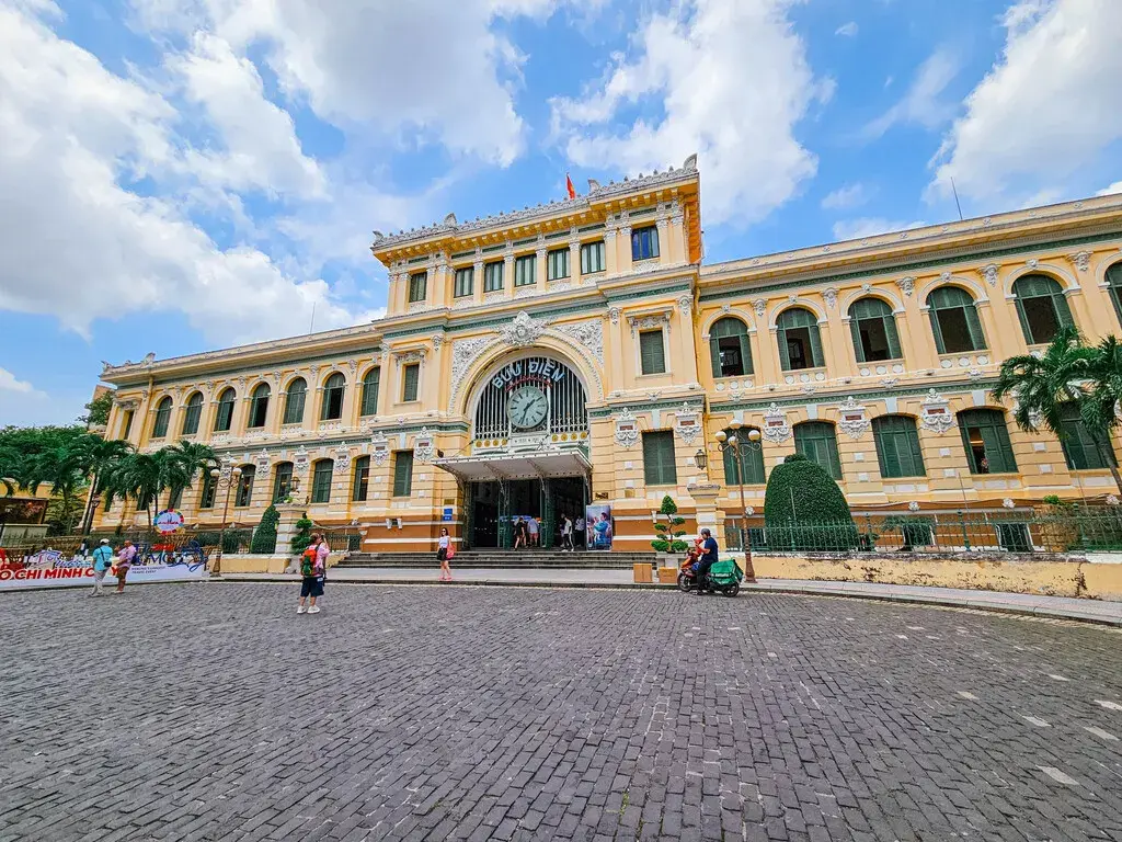 2025胡志明市旅遊指南|從行前準備到在地美食景點,一篇搞懂自由行大小事 胡志明市景點:中央郵局的正面外觀,呈現法式殖民風格建築,前方有廣場和行人。 (Front exterior of Saigon Central Post Office in Ho Chi Minh City, featuring French colonial architecture, with a plaza and pedestrians in front.)