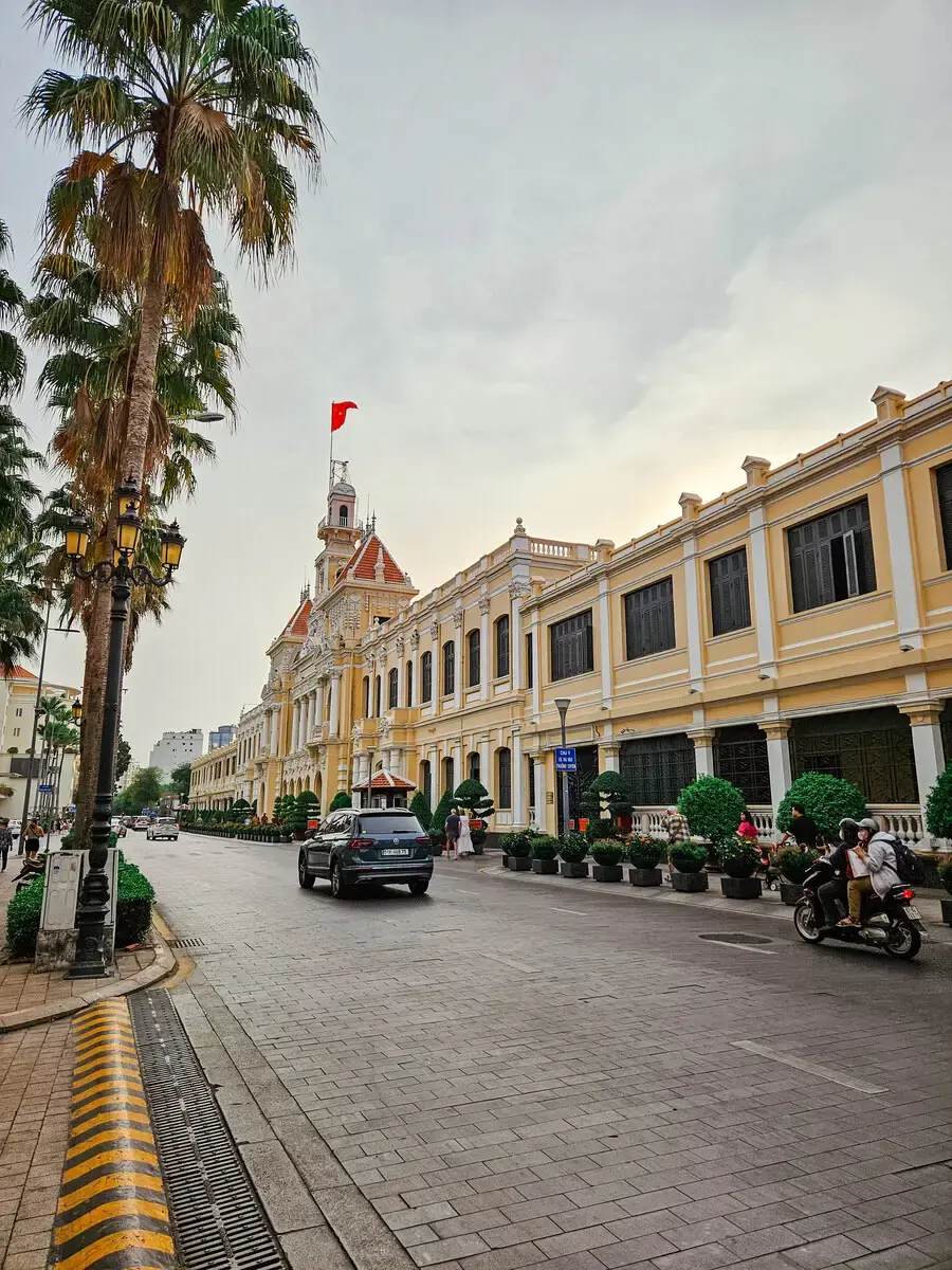 胡志明市景點-胡志明市政廳外觀，法式建築風格，前方街道有車輛及棕櫚樹(Ho Chi Minh City Hall exterior, French colonial architecture style, with vehicles and palm trees on the street in front)