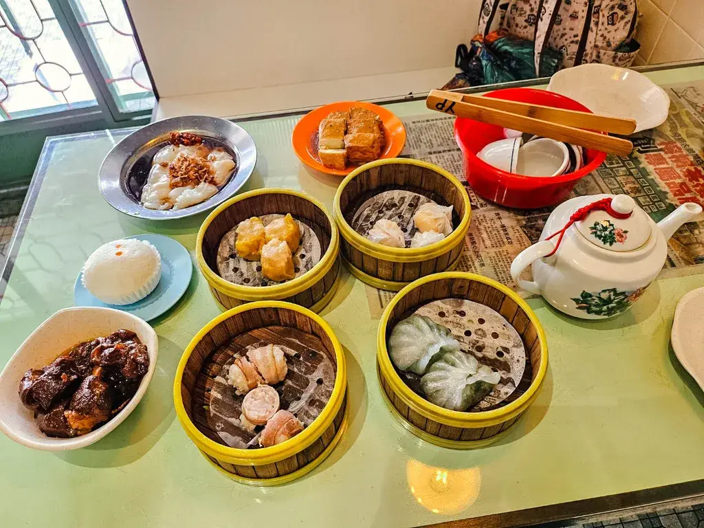 這張照片展示了豐盛的檳城美食:傳統港式點心，來自著名的榕檳茶樓。餐桌上擺滿了多籠蒸籠點心，包括蝦餃、燒賣、和腸粉、流沙包。桌面上還有一壺中國茶，呈現出在檳城享用早茶的愜意氛圍。
