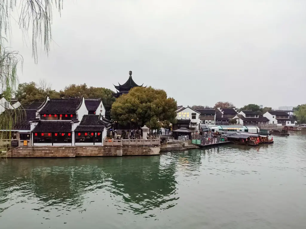 2026 蘇州旅遊攻略|5日遊深度自由行:避開人潮的園林路線、美食、住宿、交通一次搞懂 蘇州景點「七里山塘」的水鄉風貌。照片中可見白牆黑瓦的傳統江南建築、點綴其中的紅燈籠、河岸邊停泊的遊船與畫舫,以及遠處的特色飛簷古塔。山塘街是安排蘇州旅遊、蘇州自由行與規劃蘇州行程時,必訪的經典蘇州景點。