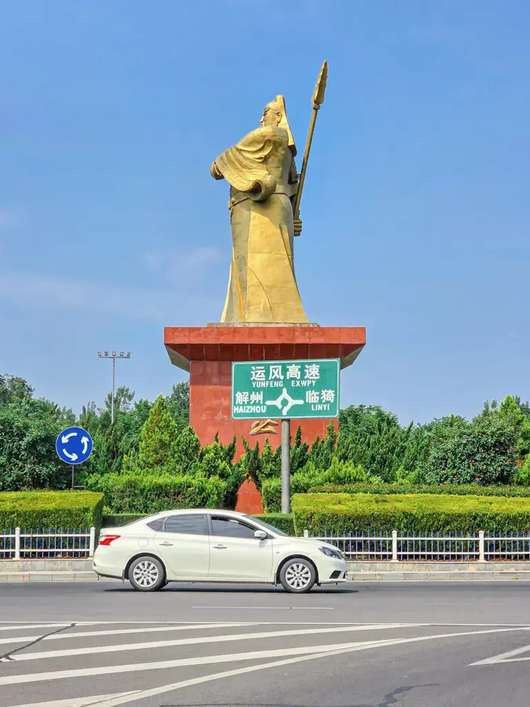 山西運城自駕前往解州關帝廟途中圓環的巨型關公金銅雕像地標,下方立有寫著「運風高速、解州、臨猗」的綠色交通指示牌,前景有一輛白色轎車在柏油公路上行駛。