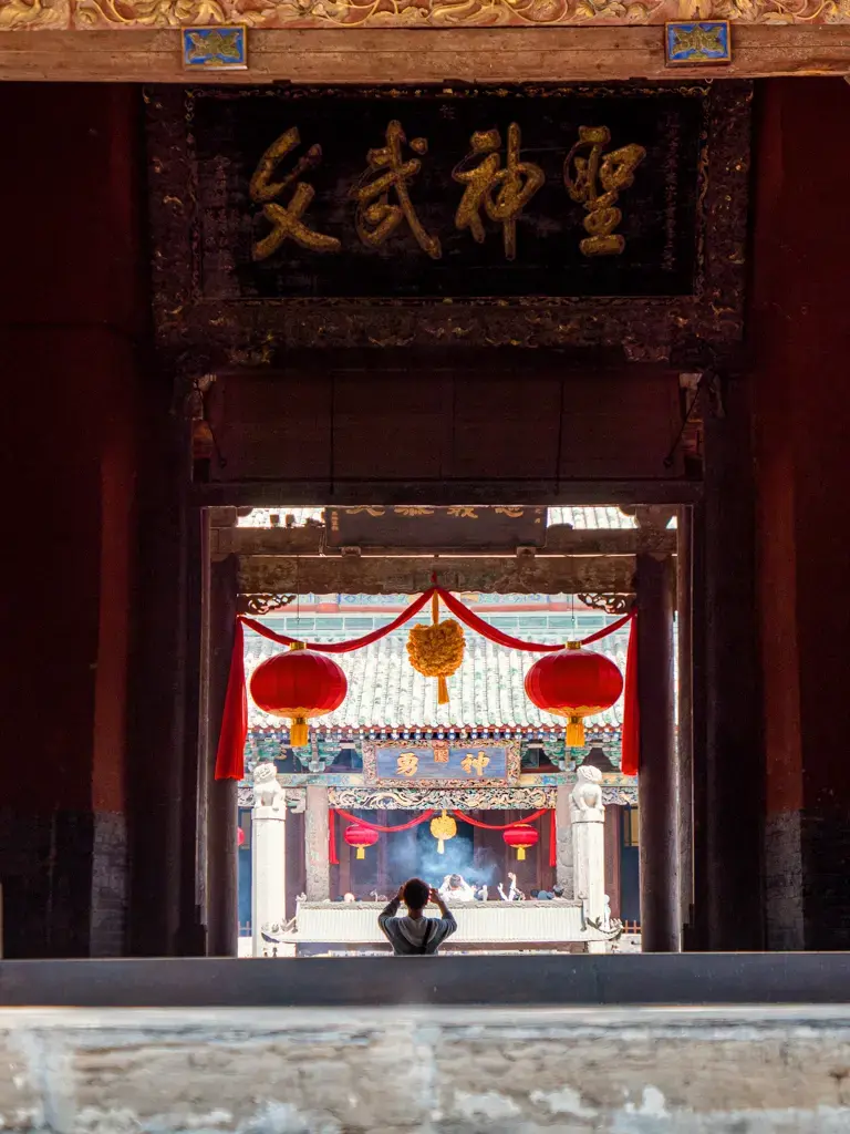 透過解州關帝廟宮殿大門對稱的門廊中軸線遠眺崇寧殿,上方懸掛著黑底金字的「聖神文武」古老匾額,前景一名自駕遊旅客正在香火裊裊的香爐前合十祈福。
