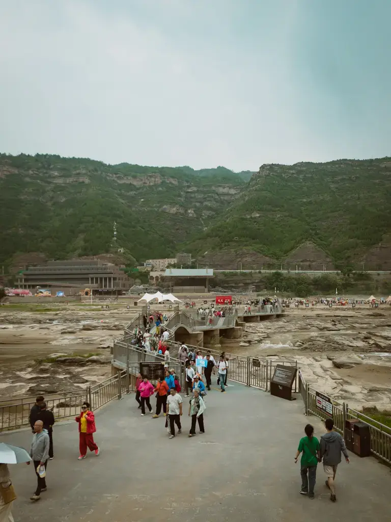 山西黃河壺口瀑布景區內的河岸觀景步道與跨河橋樑，畫面上許多自由行與自駕遊旅客正漫步於延伸至河床的灰色步道上，背景為宏偉的黃土高原斷崖與開闊的峽谷地形。