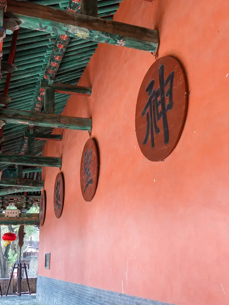 山西運城解州關帝廟內古典紅色宮牆的延伸視角,牆面掛有寫著關聖帝君封號的圓形木質字匾,上方襯托著帶有精緻彩繪的傳統中式木構屋簷與斗拱結構。