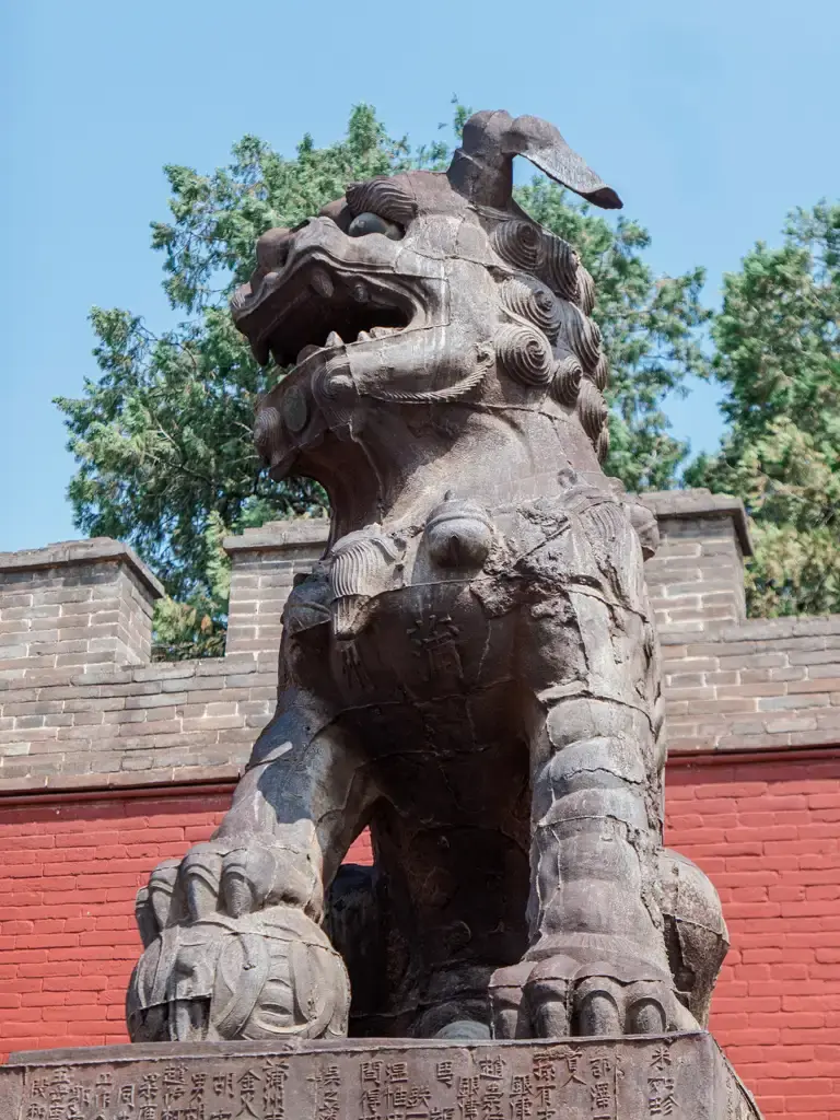 解州關帝廟內珍藏的古老雕刻獅子塑像近景特寫,刻工粗獷且帶有歲月斑駁痕跡,下方的石碑底座刻滿了歷史銘文,背景為傳統的紅牆與青磚城垛。