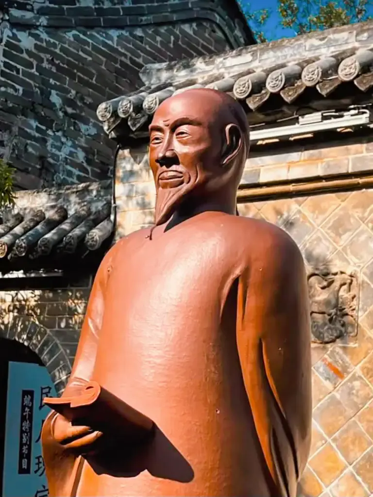 陝西韓城古城歷史名士文化景點實拍:一尊手持書卷的歷史人物(清代重臣:王杰)棕褐色雕像立於古樸的青磚黛瓦與雕花照壁前,展現古城深厚的文史底蘊。